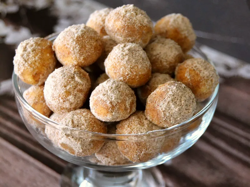 A glass bowl heaped with golden cinnamon-sugar coated donut holes on a dark wooden table