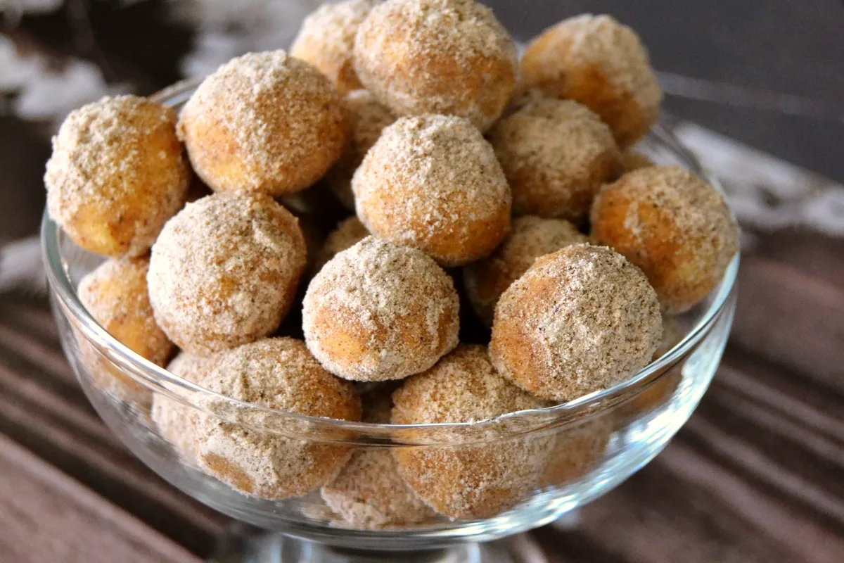 A glass bowl heaped with golden cinnamon-sugar coated donut holes on a dark wooden table