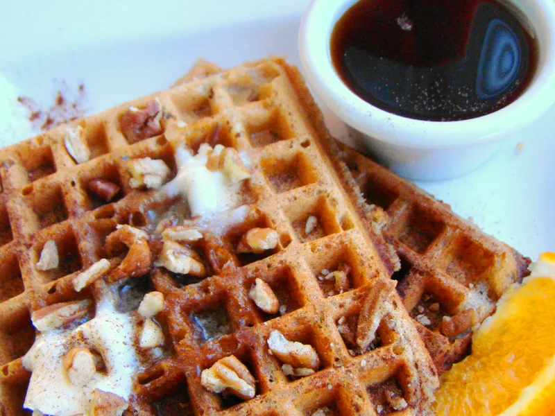 Golden pumpkin waffles topped with butter and chopped pecans, with a cup of maple syrup and orange slices