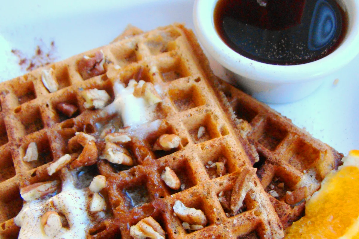 Golden pumpkin waffles topped with butter and chopped pecans, with a cup of maple syrup and orange slices