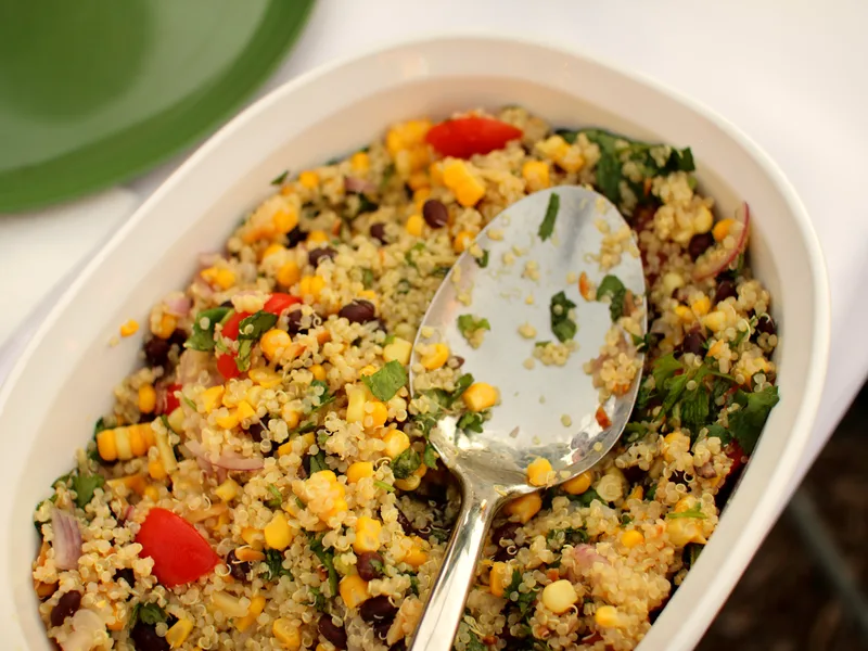Oval dish of quinoa salad with sweet corn, black beans, cherry tomatoes, red onion and chopped cilantro with a serving spoon