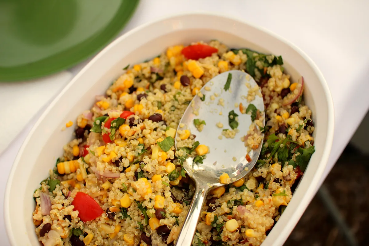 Oval dish of quinoa salad with sweet corn, black beans, cherry tomatoes, red onion and chopped cilantro with a serving spoon