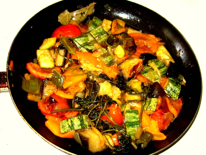 A black skillet seen from above, filled with chunky ratatouille - striped zucchini pieces, eggplant, red and orange peppers, tomato and bay leaves, all glossy with olive oil