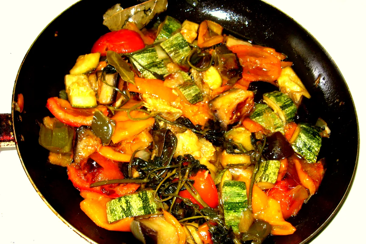 A black skillet seen from above, filled with chunky ratatouille - striped zucchini pieces, eggplant, red and orange peppers, tomato and bay leaves, all glossy with olive oil