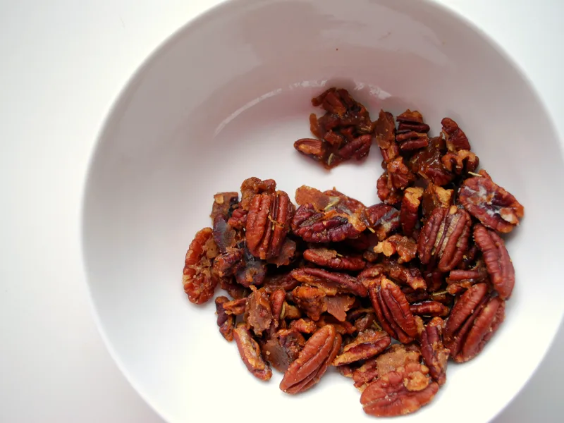 Glossy candied pecan halves flecked with rosemary in a white bowl