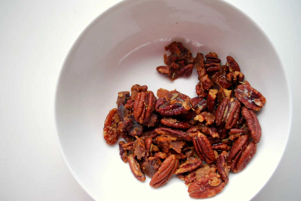 Glossy candied pecan halves flecked with rosemary in a white bowl