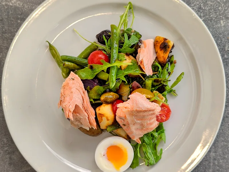 A white plate of salmon nicoise salad seen from above with large flakes of cooked salmon, green beans, cherry tomatoes, roasted potato chunks, green and black olives and arugula, with a halved soft-boiled egg with a bright orange yolk in front