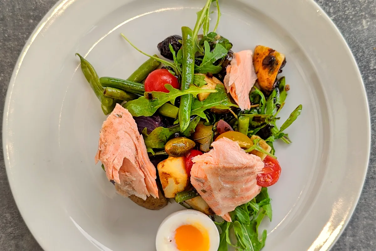 A white plate of salmon nicoise salad seen from above with large flakes of cooked salmon, green beans, cherry tomatoes, roasted potato chunks, green and black olives and arugula, with a halved soft-boiled egg with a bright orange yolk in front
