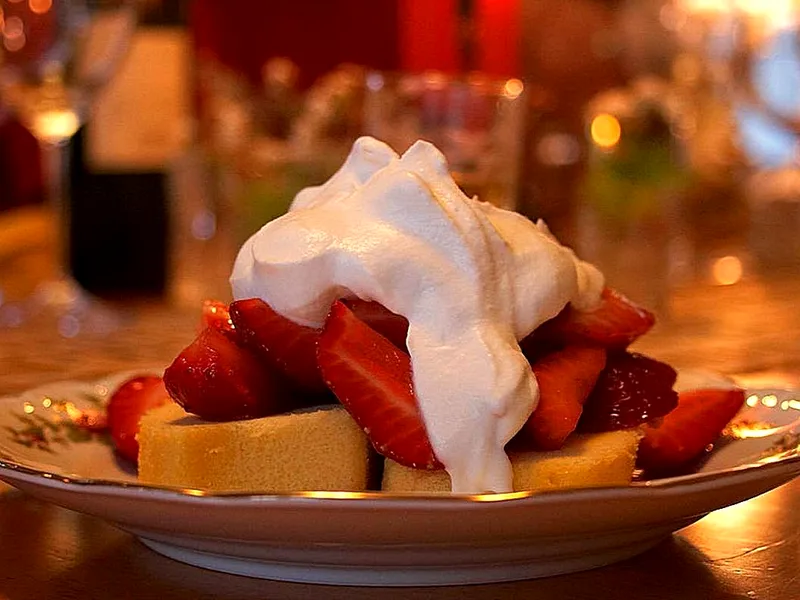 Slices of pale sponge cake on a small plate piled with sliced macerated strawberries and a large mound of softly whipped cream draped over the top, in warm candlelight