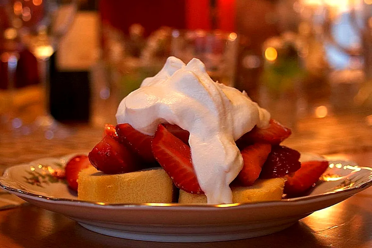 Slices of pale sponge cake on a small plate piled with sliced macerated strawberries and a large mound of softly whipped cream draped over the top, in warm candlelight