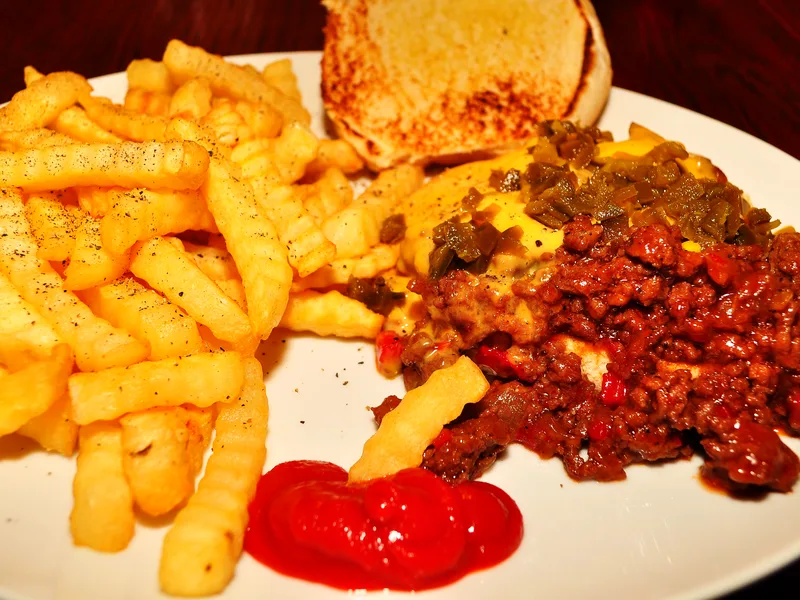 Open-face sloppy joe on a toasted bun topped with melted cheese and chopped pickled peppers, served on a white plate with a pile of peppered crinkle-cut fries and a dollop of ketchup