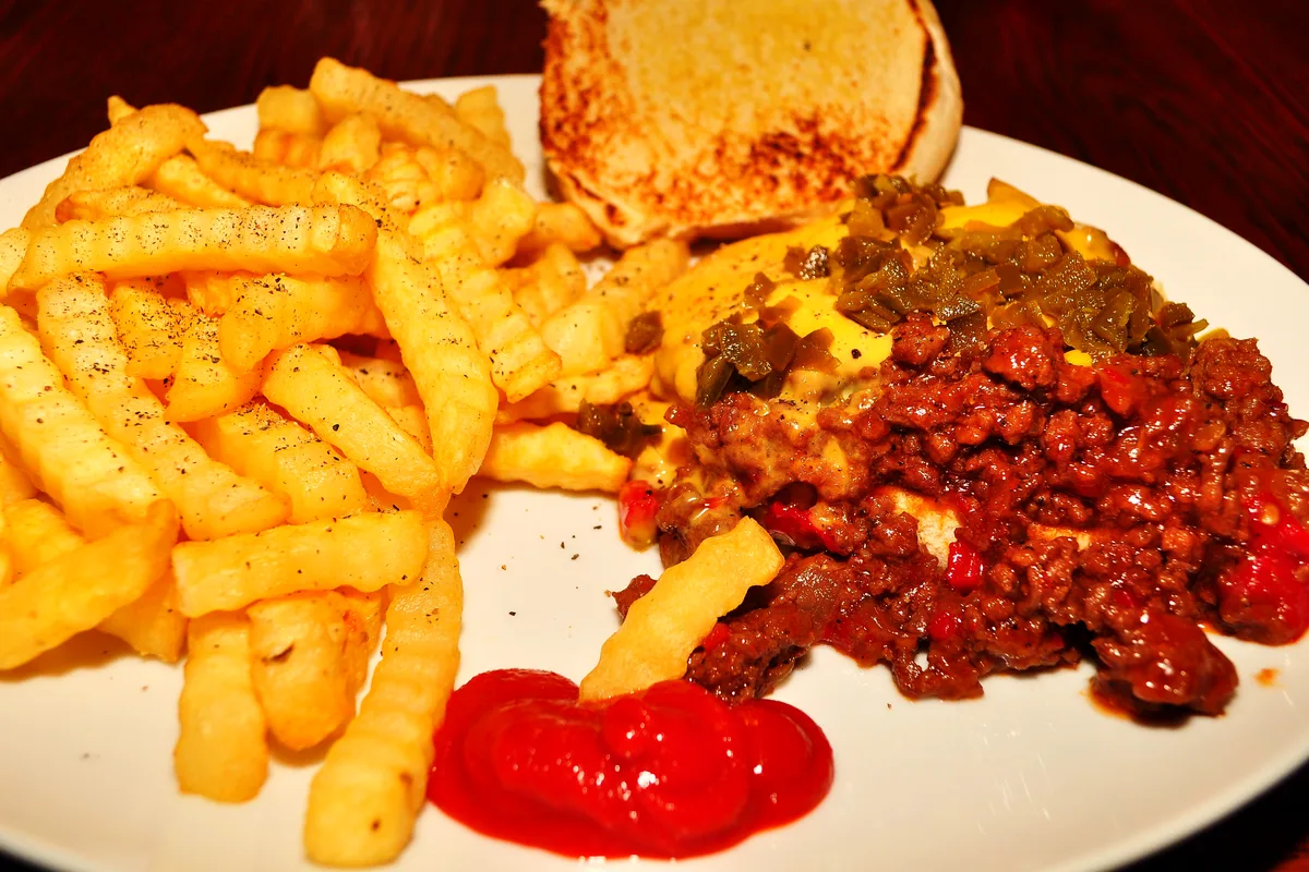Open-face sloppy joe on a toasted bun topped with melted cheese and chopped pickled peppers, served on a white plate with a pile of peppered crinkle-cut fries and a dollop of ketchup