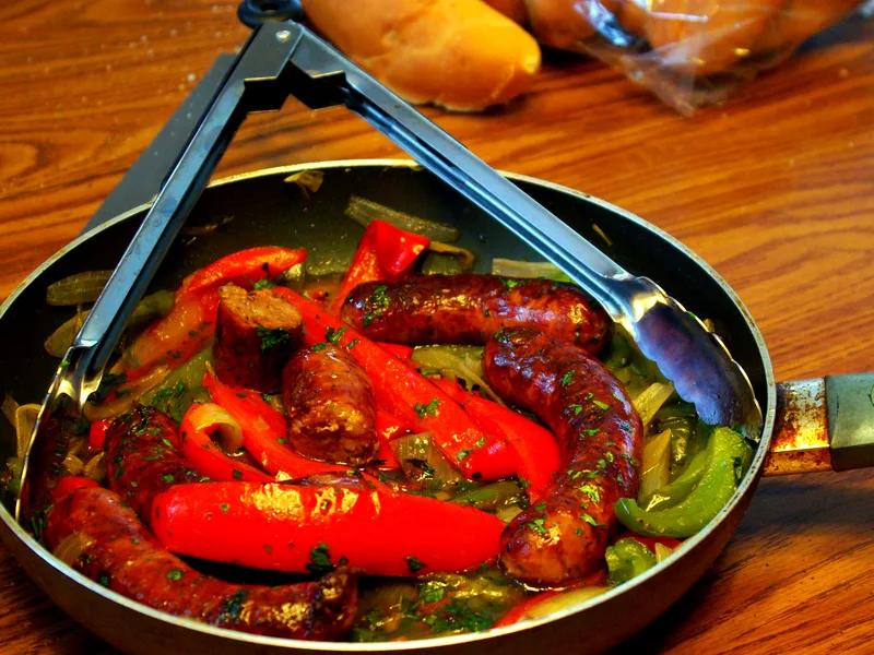 Skillet of browned Italian sausages with sliced red and green peppers and onions, scattered with fresh parsley
