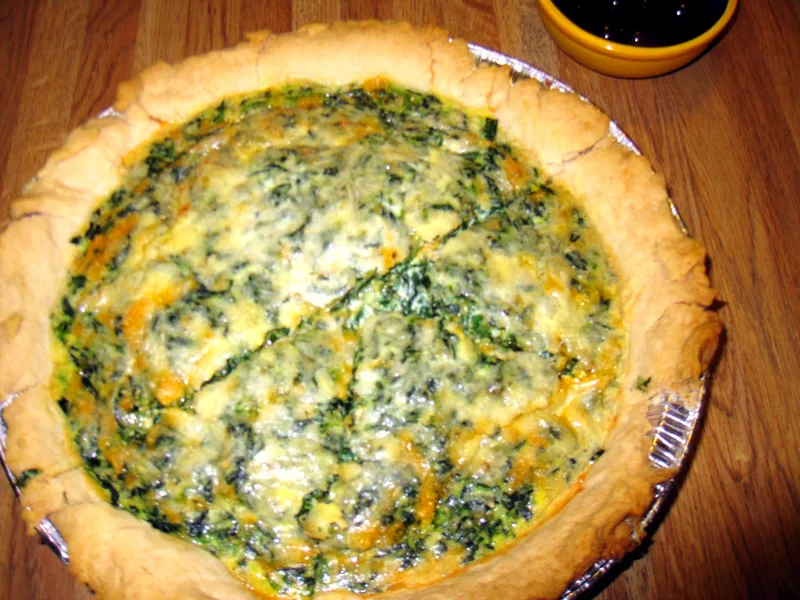 A whole spinach quiche in a pie tin with a rustic golden crust and a spinach and cheese filling flecked with two cheeses