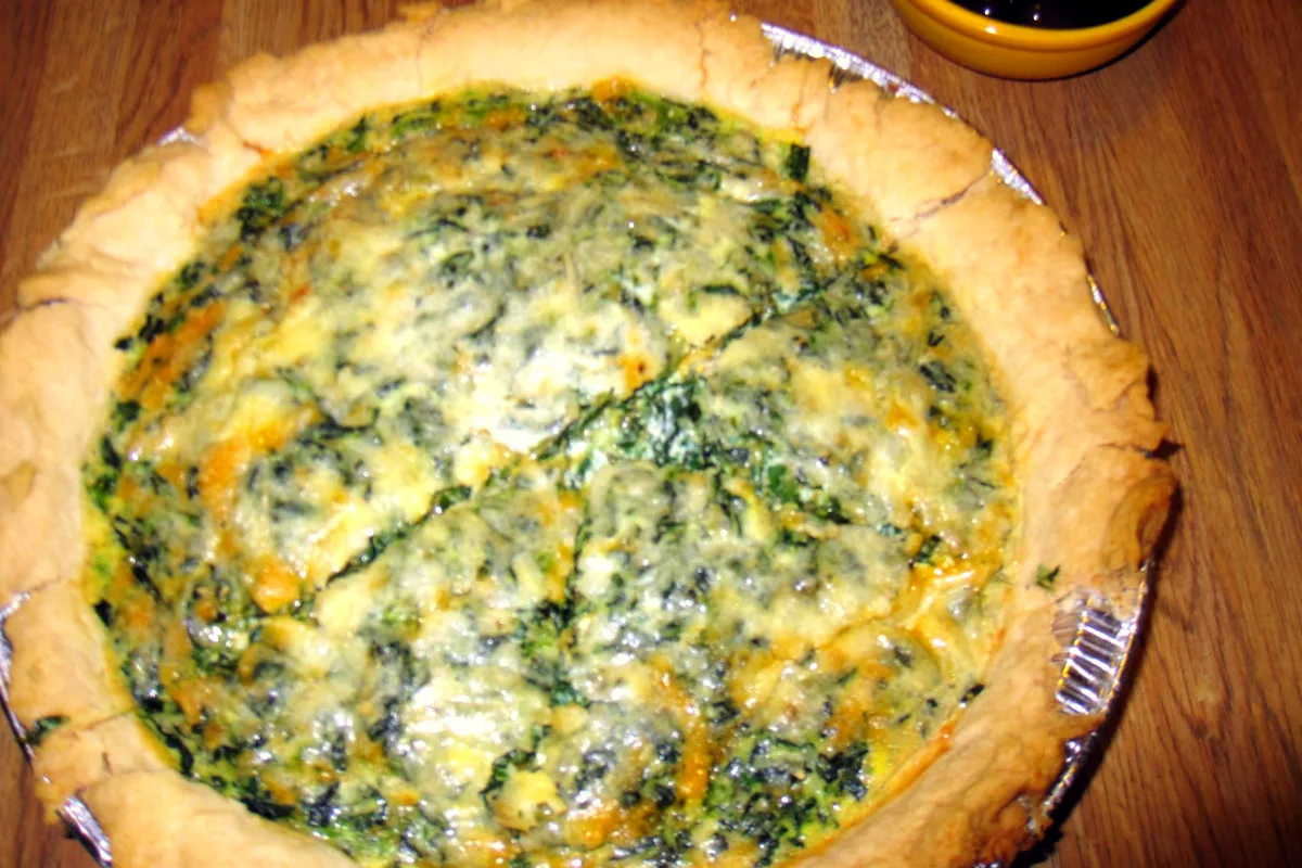 A whole spinach quiche in a pie tin with a rustic golden crust and a spinach and cheese filling flecked with two cheeses