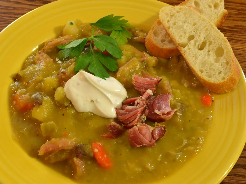 Bowl of slow cooker split pea soup with shredded ham, carrots and a dollop of sour cream, with crusty bread on the side