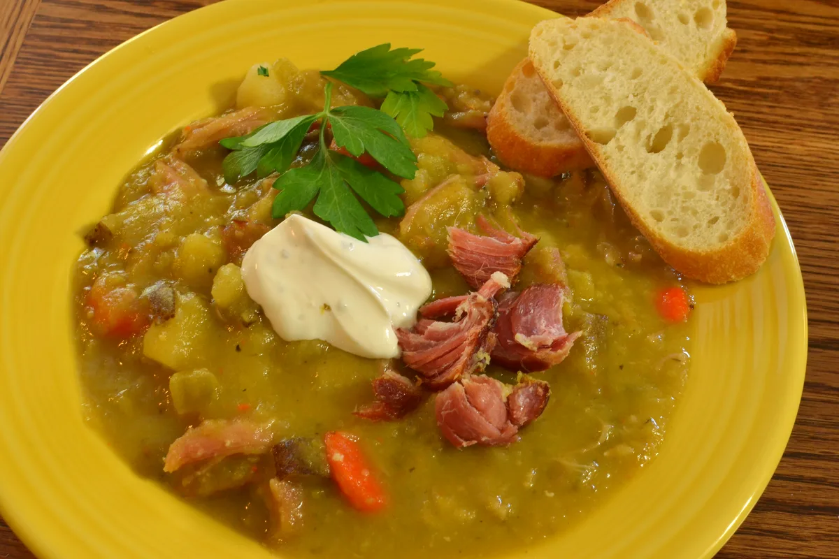 Bowl of slow cooker split pea soup with shredded ham, carrots and a dollop of sour cream, with crusty bread on the side