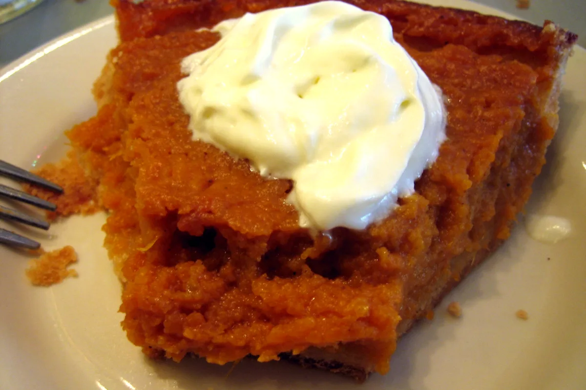 A slice of classic sweet potato pie topped with a swirl of whipped cream on a white plate