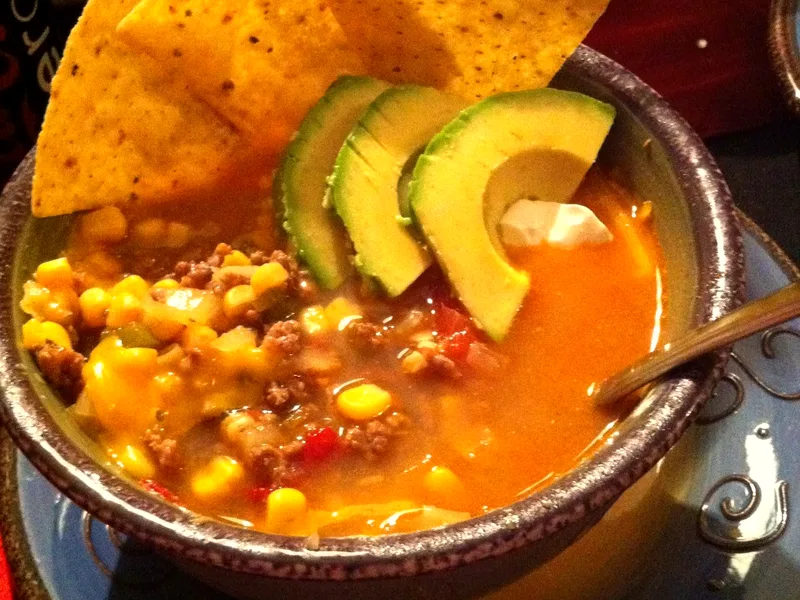 A rustic bowl of taco soup with ground beef, corn and melted cheese, topped with avocado slices and tortilla chips and a spoonful of sour cream