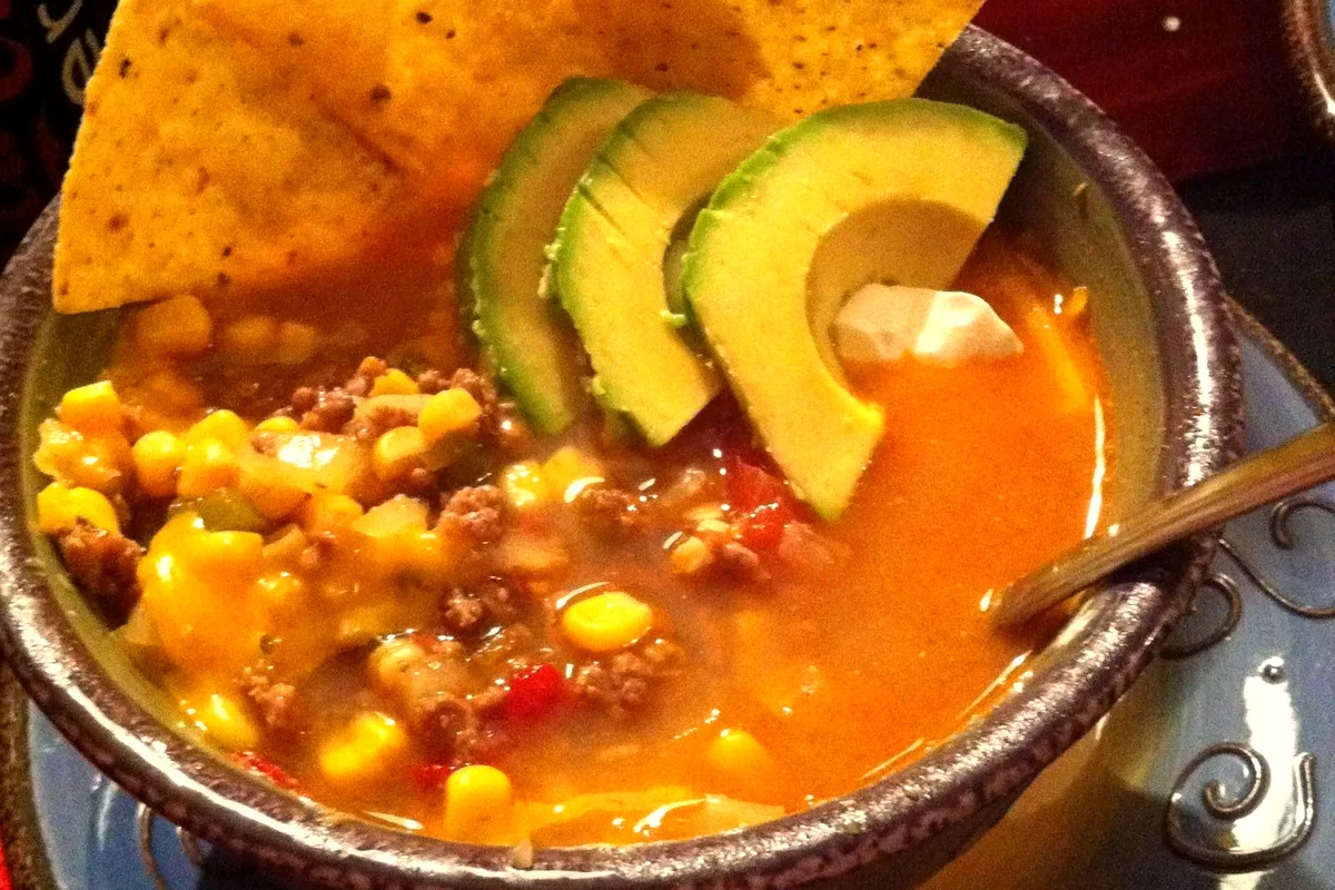 A rustic bowl of taco soup with ground beef, corn and melted cheese, topped with avocado slices and tortilla chips and a spoonful of sour cream