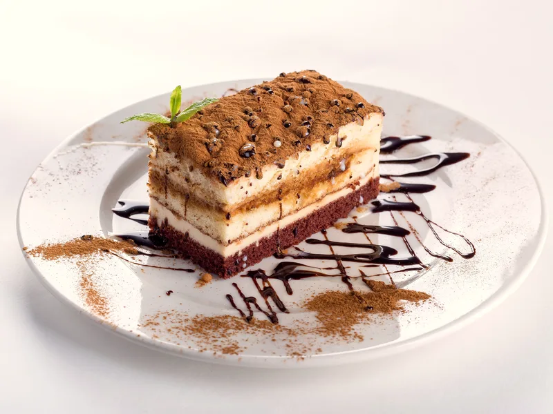 Slice of classic tiramisu with cocoa-dusted top, pale mascarpone layers and a dark sponge base, chocolate sauce on the plate