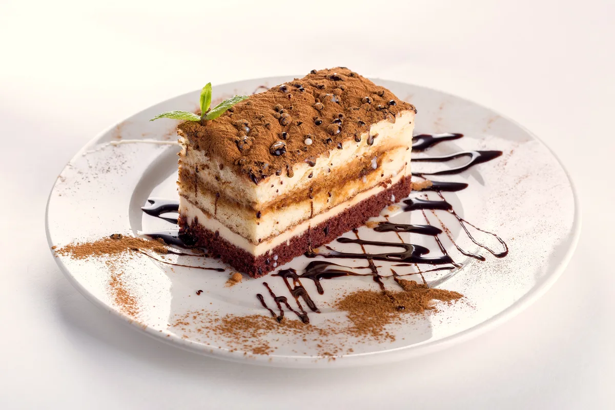 Slice of classic tiramisu with cocoa-dusted top, pale mascarpone layers and a dark sponge base, chocolate sauce on the plate