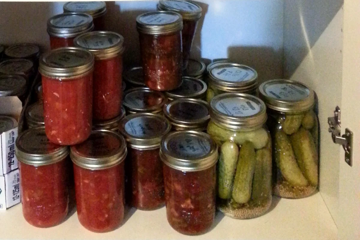 Home-canned jars of red tomato sauce stacked on a pantry shelf alongside other preserved jars, the cropped view centred on the tomato sauce jars