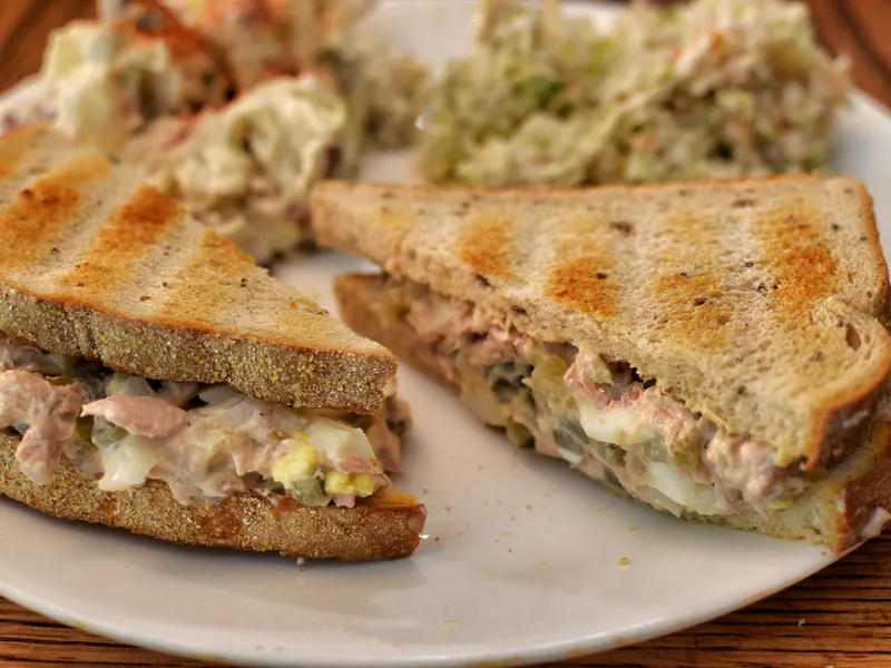 Tuna salad sandwich on toasted rye bread, cut in half, with creamy tuna and egg filling spilling out, coleslaw behind