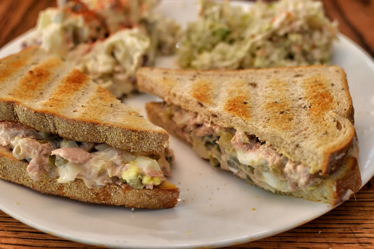 Tuna salad sandwich on toasted rye bread, cut in half, with creamy tuna and egg filling spilling out, coleslaw behind