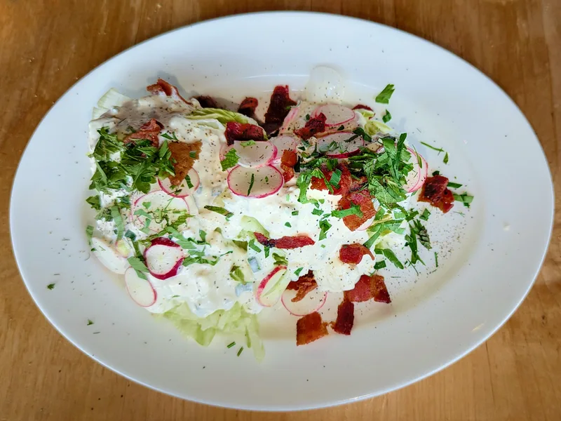Two iceberg lettuce wedges on a white oval plate covered with creamy blue cheese dressing, topped with crisp bacon pieces, thinly sliced radishes, chopped parsley and black pepper