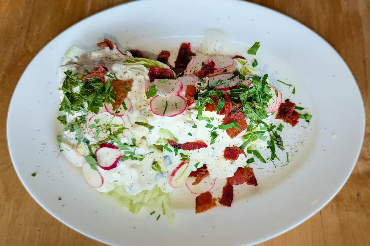 Two iceberg lettuce wedges on a white oval plate covered with creamy blue cheese dressing, topped with crisp bacon pieces, thinly sliced radishes, chopped parsley and black pepper