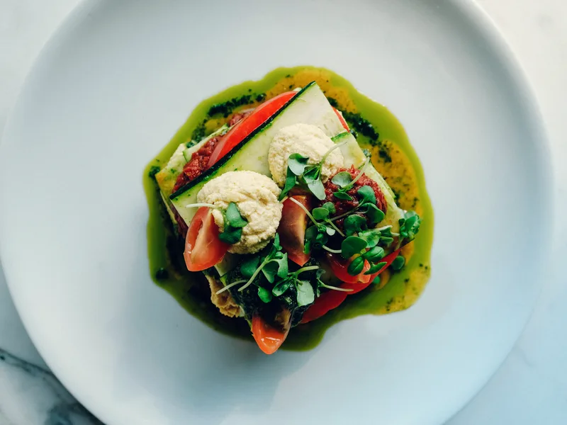 Stack of raw zucchini lasagna with cashew cream, tomato sauce and basil pesto on a white plate, topped with microgreens