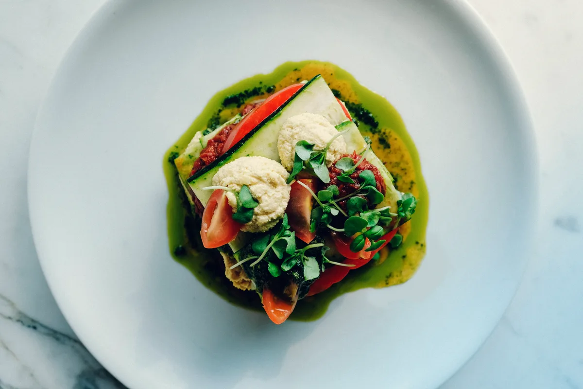 Stack of raw zucchini lasagna with cashew cream, tomato sauce and basil pesto on a white plate, topped with microgreens